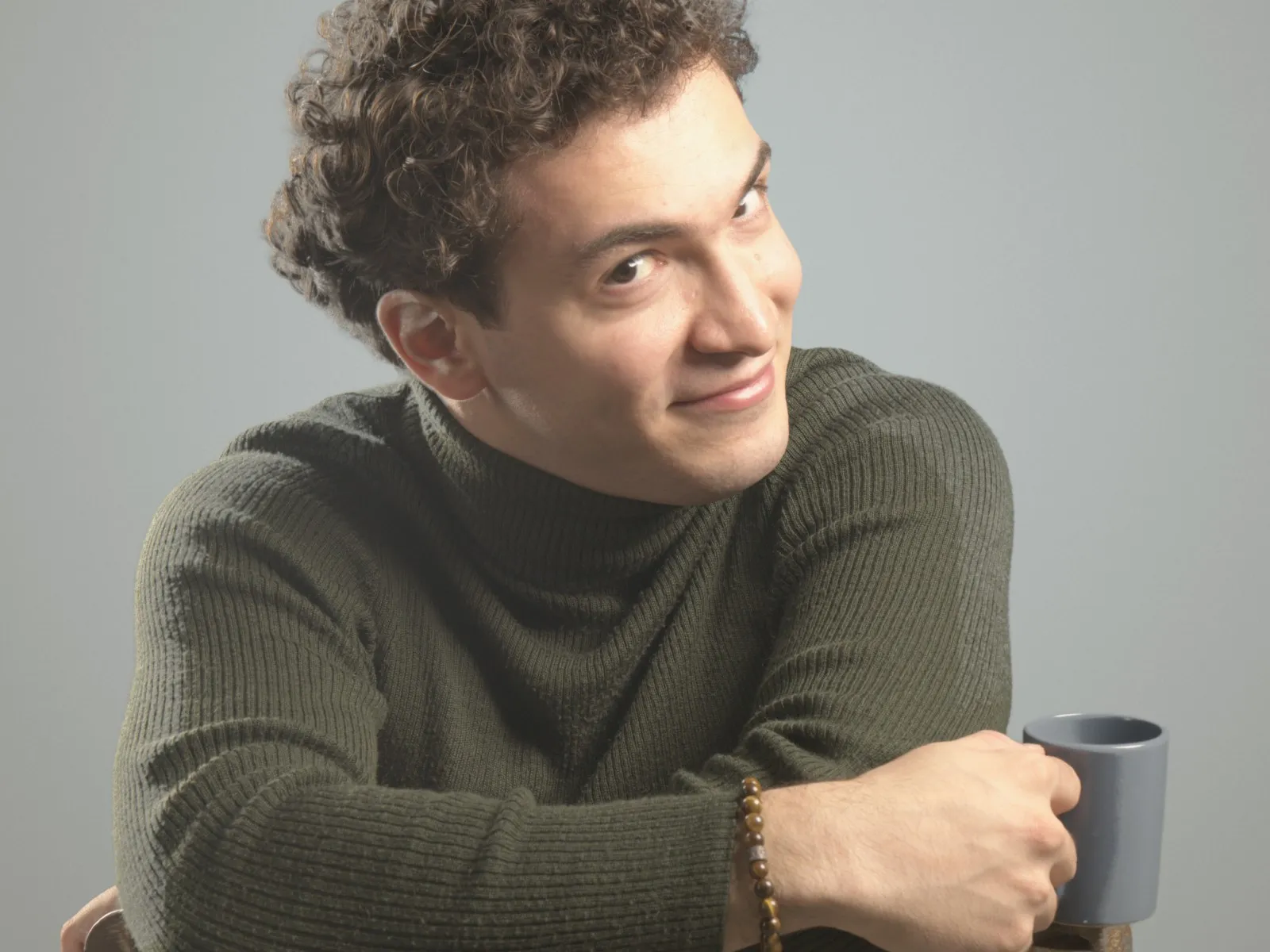 Baritone Biran Özgür Sariyer, studio portrait, with a cup, leaning on a chair, smiling